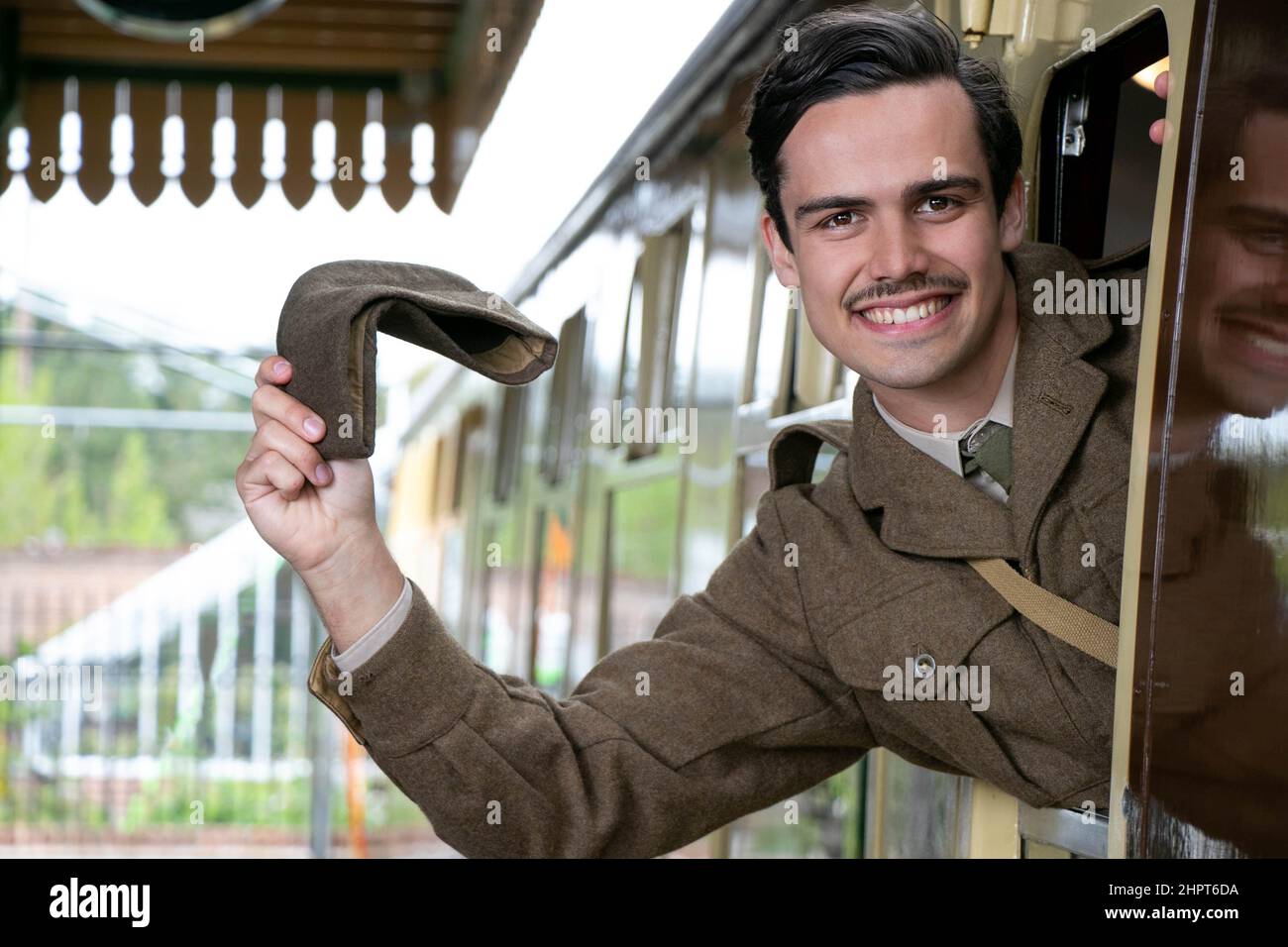 Attractive male british soldier in vintage WW2 uniform at train station, leaning out of train window, waving, smiling. Stock Photo
