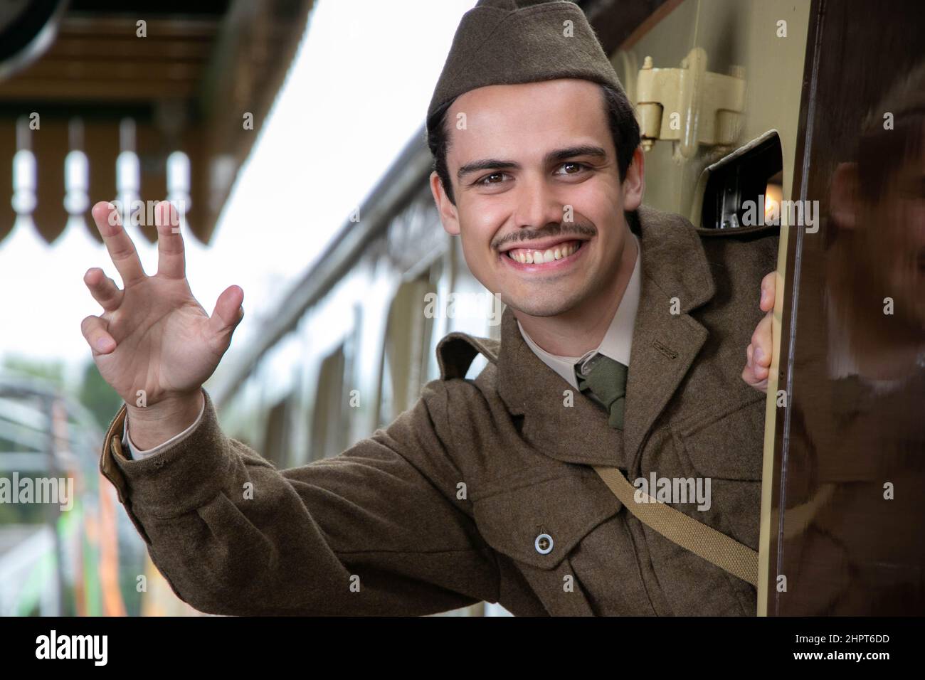 Attractive male british soldier in vintage WW2 uniform at train station, leaning out of train, waving and smiling Stock Photo