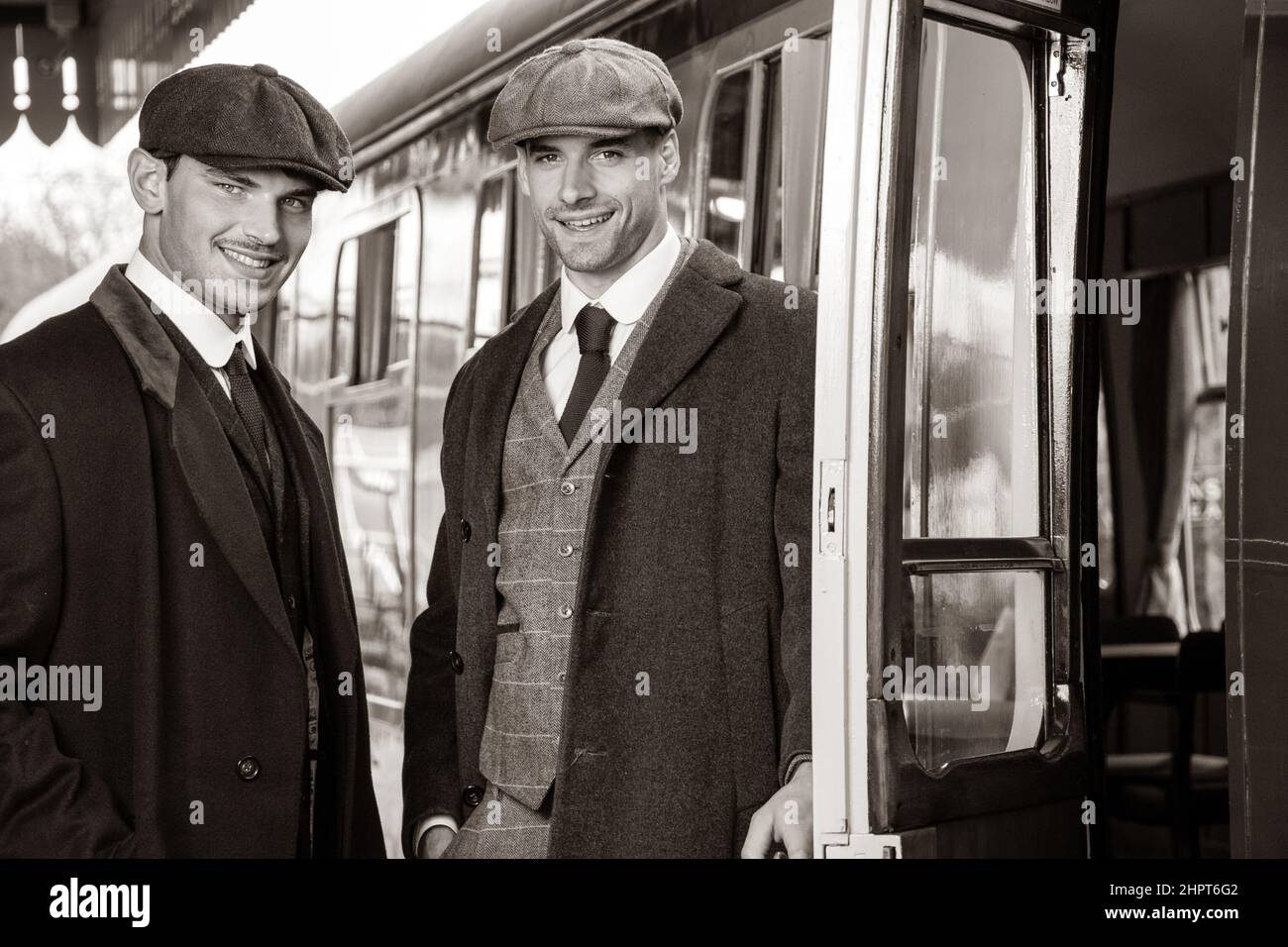 Attractive English gangsters standing next to train at railway station, laughing, with train in the background Stock Photo