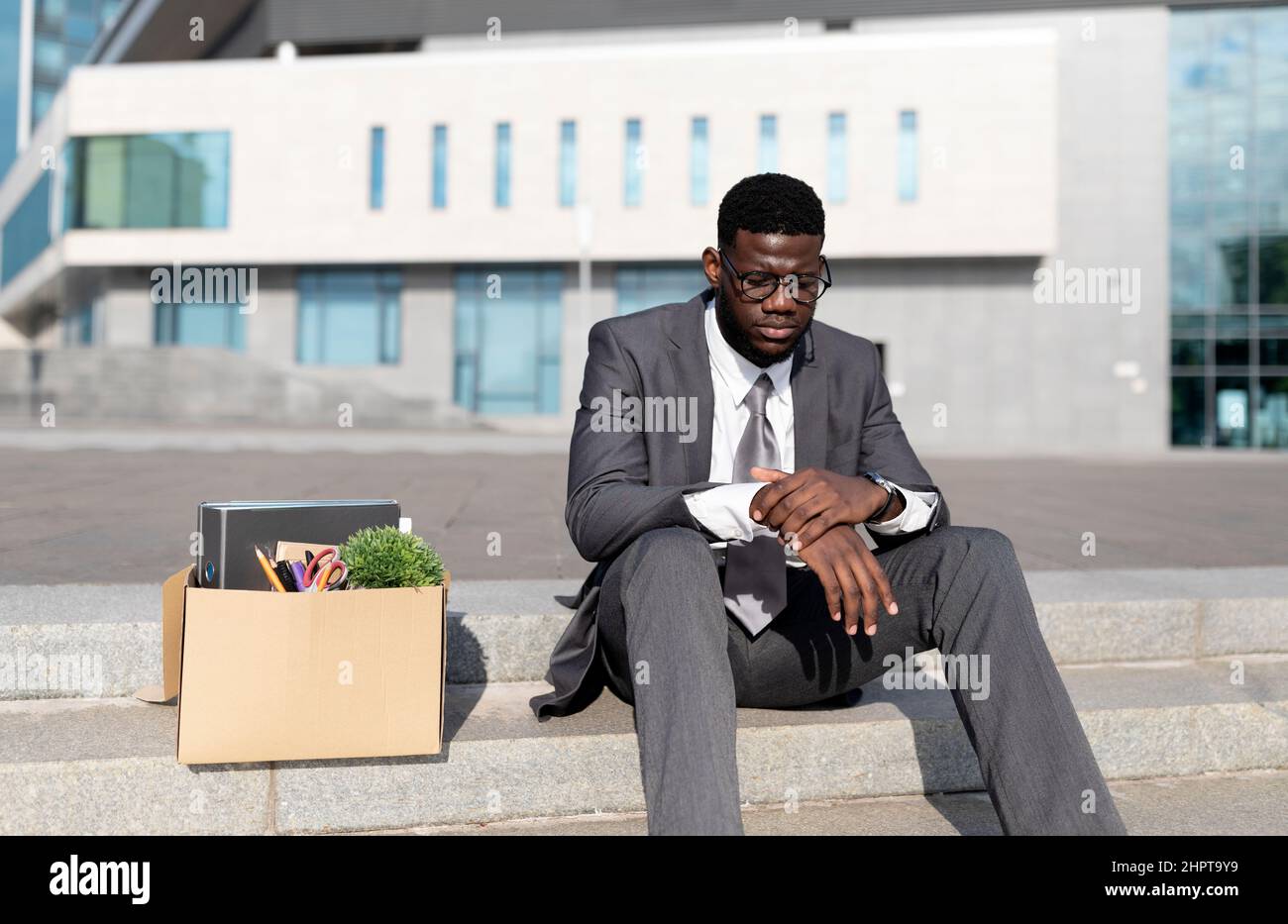 Crisis and unemployment concept. Upset black office worker sitting with box of personal stuff on stairs of office center Stock Photo