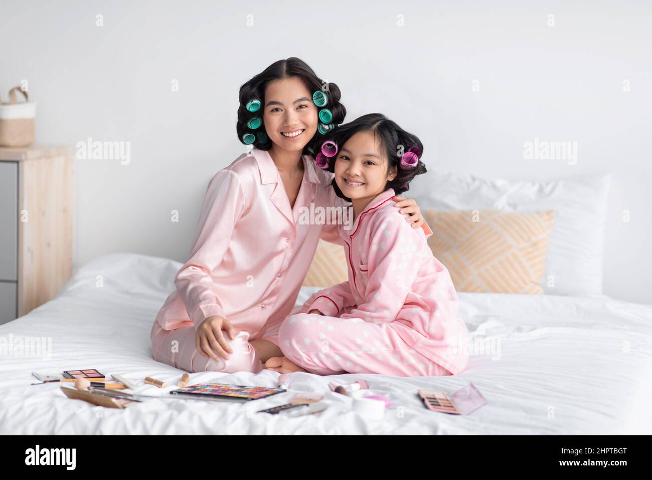 Satisfied young japanese female and teen girl in pajamas and curlers with cosmetics on bed hugs in bedroom Stock Photo