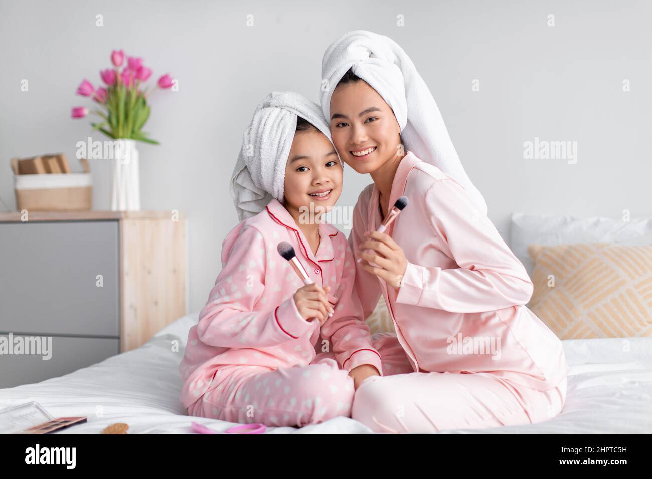 Smiling young pretty japanese female and teenage girl in pajamas and towel sit on bed with makeup brushes Stock Photo
