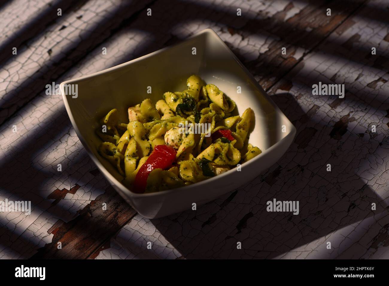 Mozzarella, tomato and pesto pasta salad in a white square dish on a distressed table with sun window shadows falling across the table and dish. Stock Photo