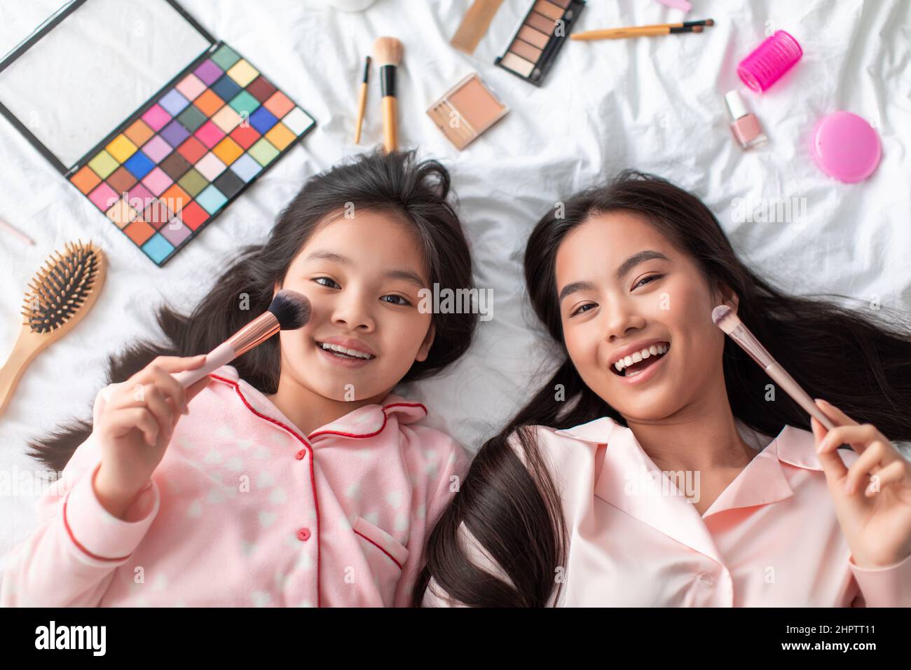 Cheerful young japanese female and teenage girl in pajamas lie with cosmetics on bed, have fun Stock Photo