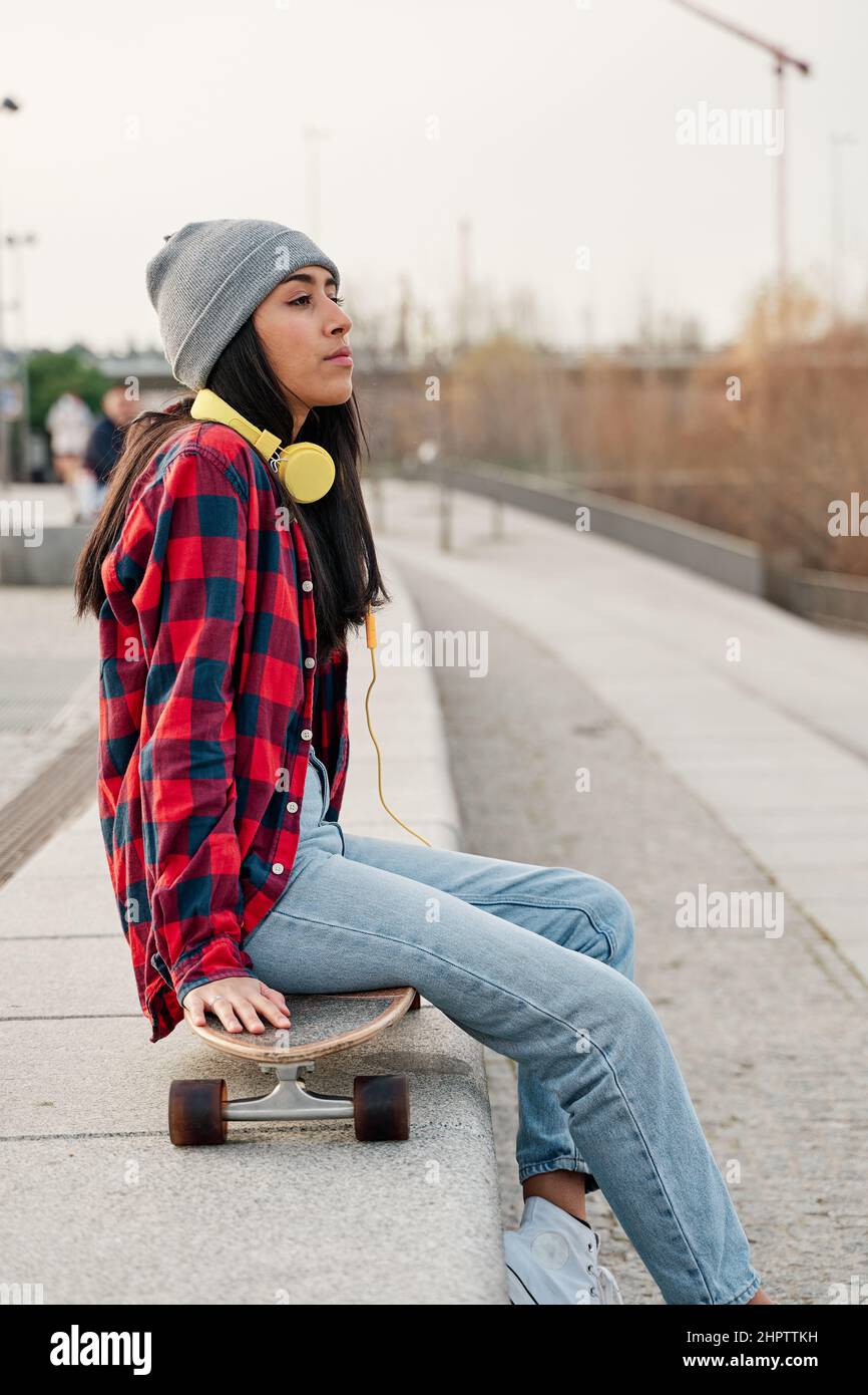 Young Hispanic woman sitting with skateboard on her knees. Venezuelan Latin woman with headphones. Stock Photo