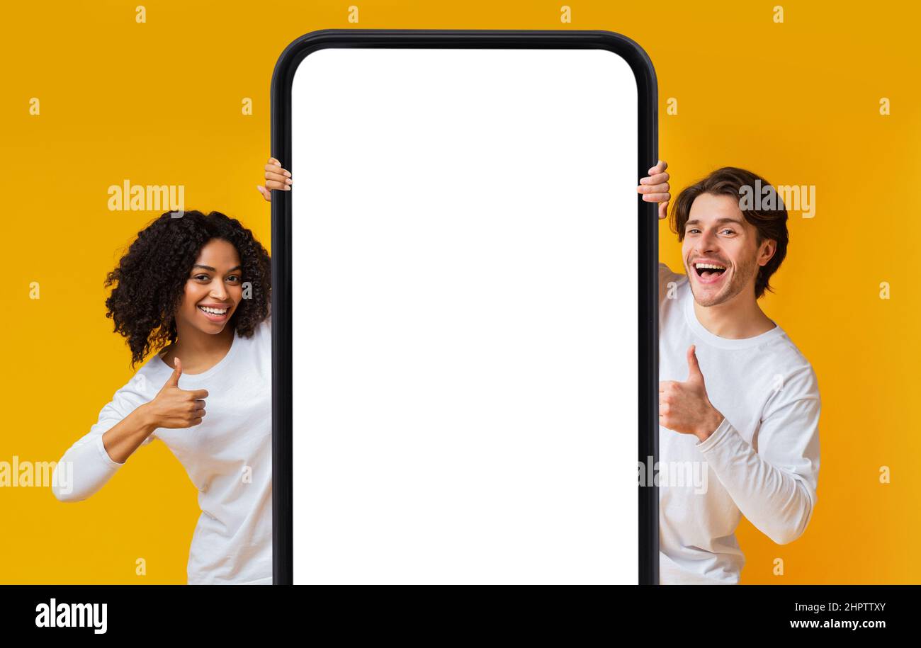 Cheerful interracial couple showing cellphone with blank screen, gesturing thumbs up, recommending mobile app, mockup Stock Photo