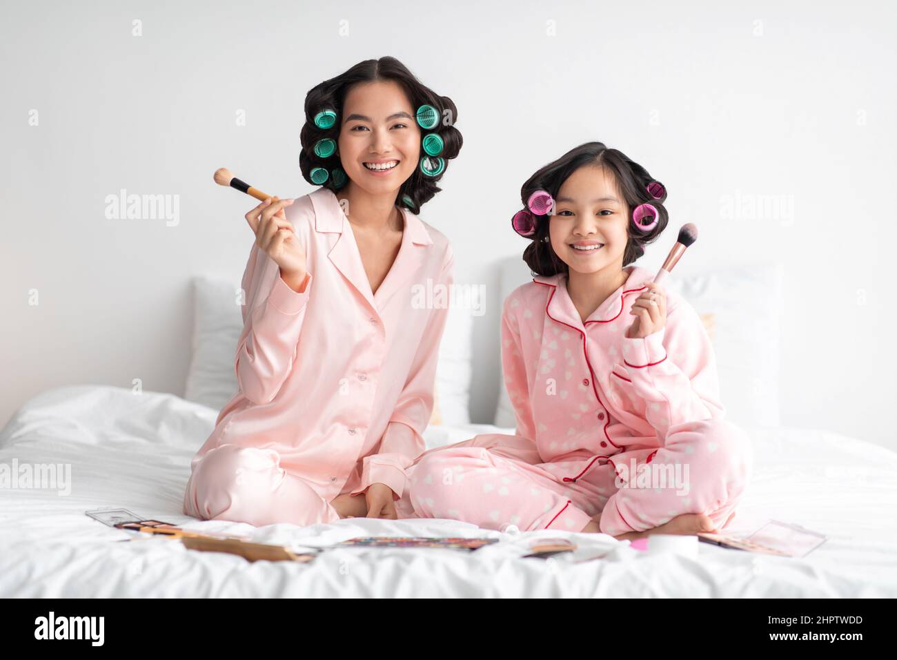 Satisfied young japanese female and teenage girl in pajamas and curlers with makeup brushes on bed Stock Photo