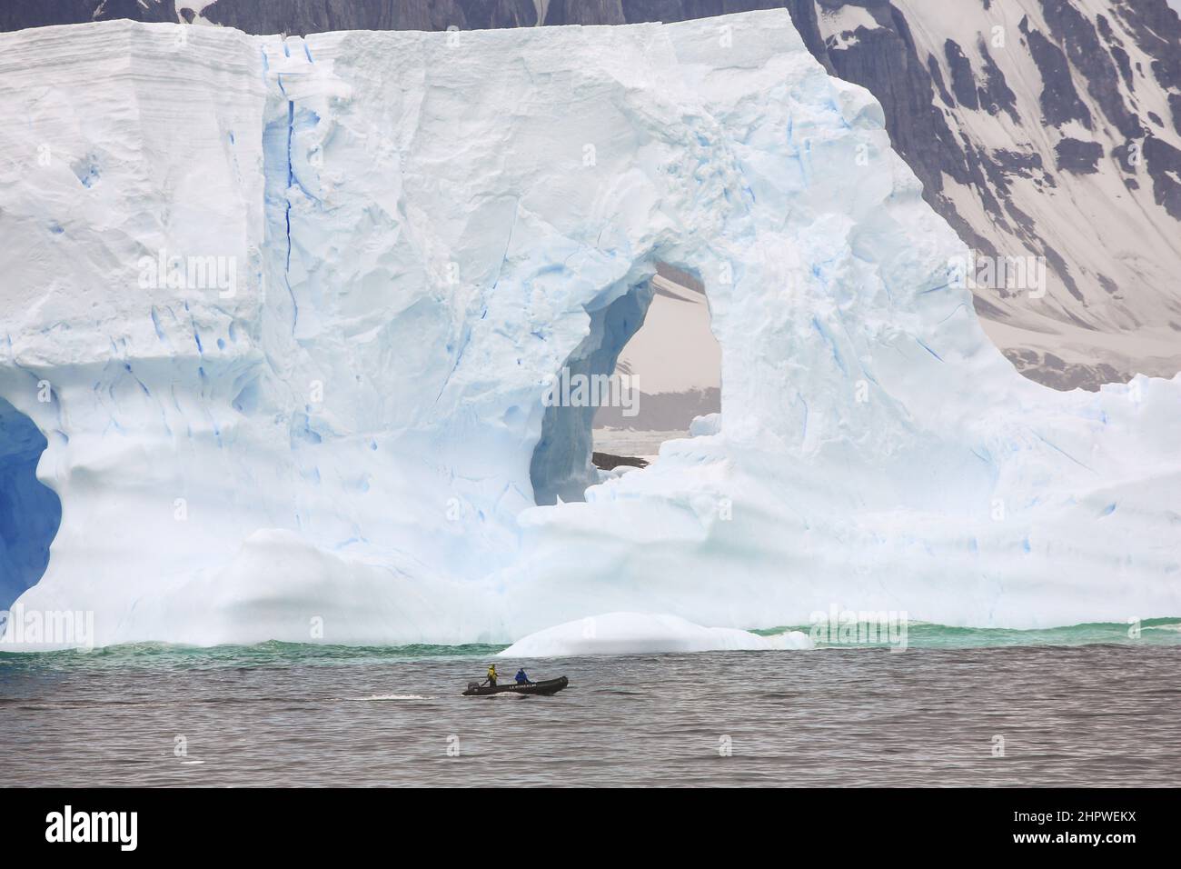 Two crew in a zodiac boat from Le Boreal cruise ship pass a huge iceberg in the South Atlantic Ocean, south-west of Petermann Island, Antarctica. Stock Photo