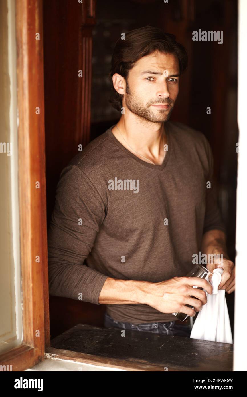 Long night ahead. Shot of a handsome young man drying off a glass while looking through a window. Stock Photo