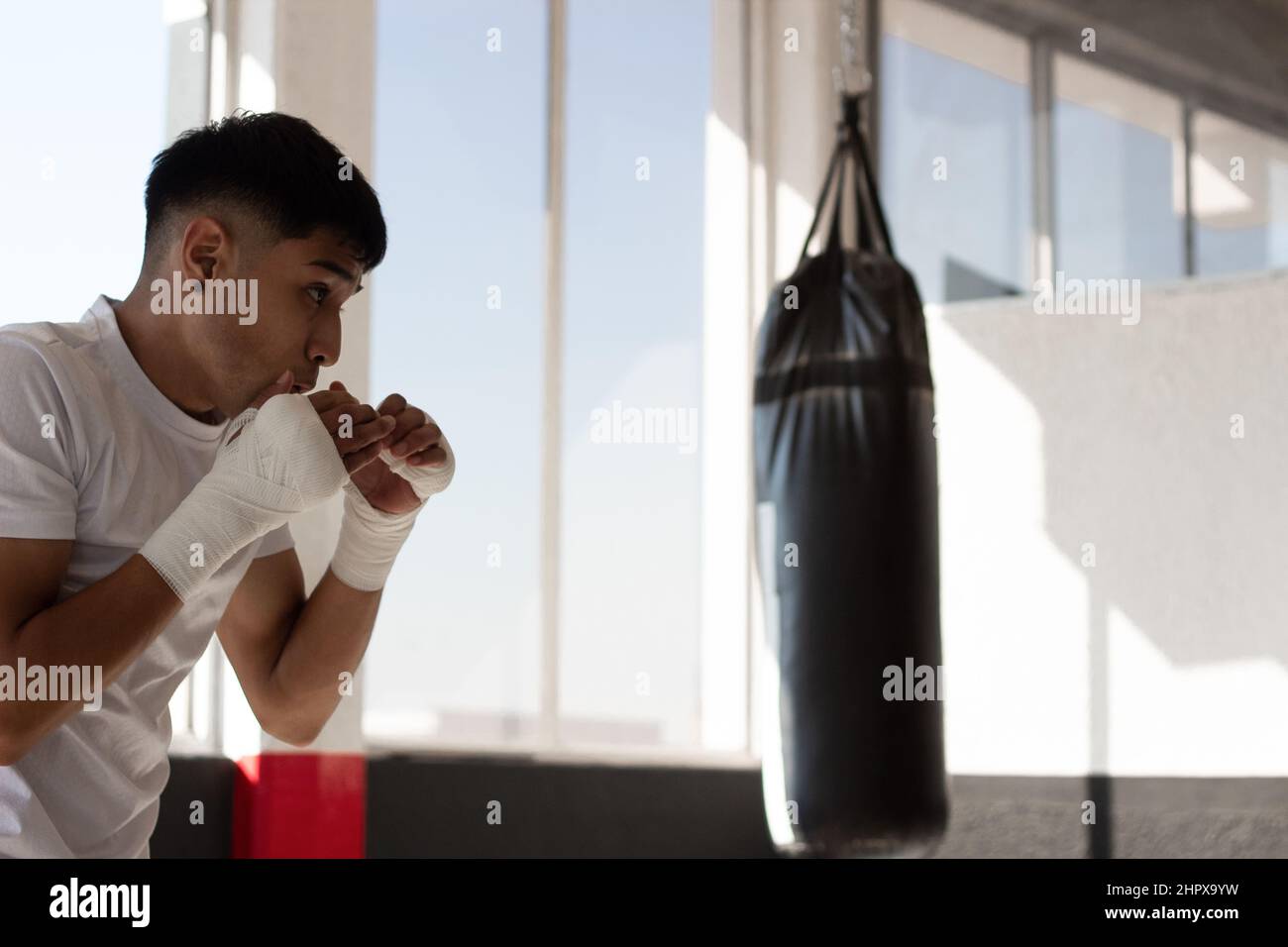 young boxer wearing a white t-shirt shadow boxing with his fists bandaged during his training inside the boxing gym. Stock Photo