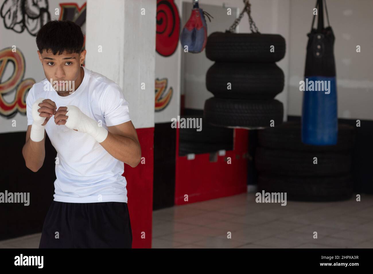 young boxer wearing a white t-shirt shadow boxing with his fists bandaged during his training inside the boxing gym. Stock Photo