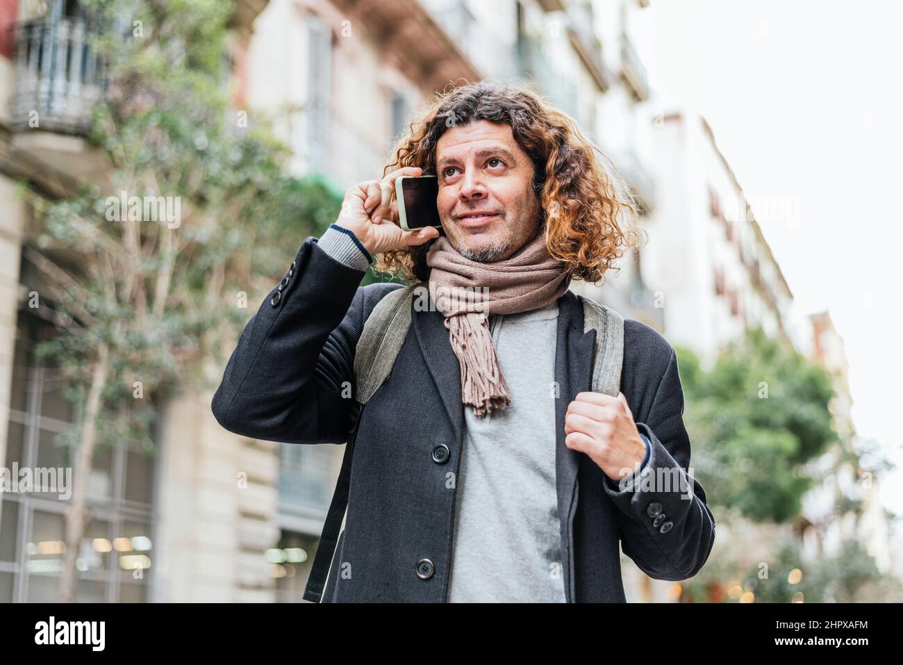 Low angle of positive mature male in smart casual clothes with backpack and scarf. He is looking away with smile and speaking on cellphone on blurred background of city street Stock Photo