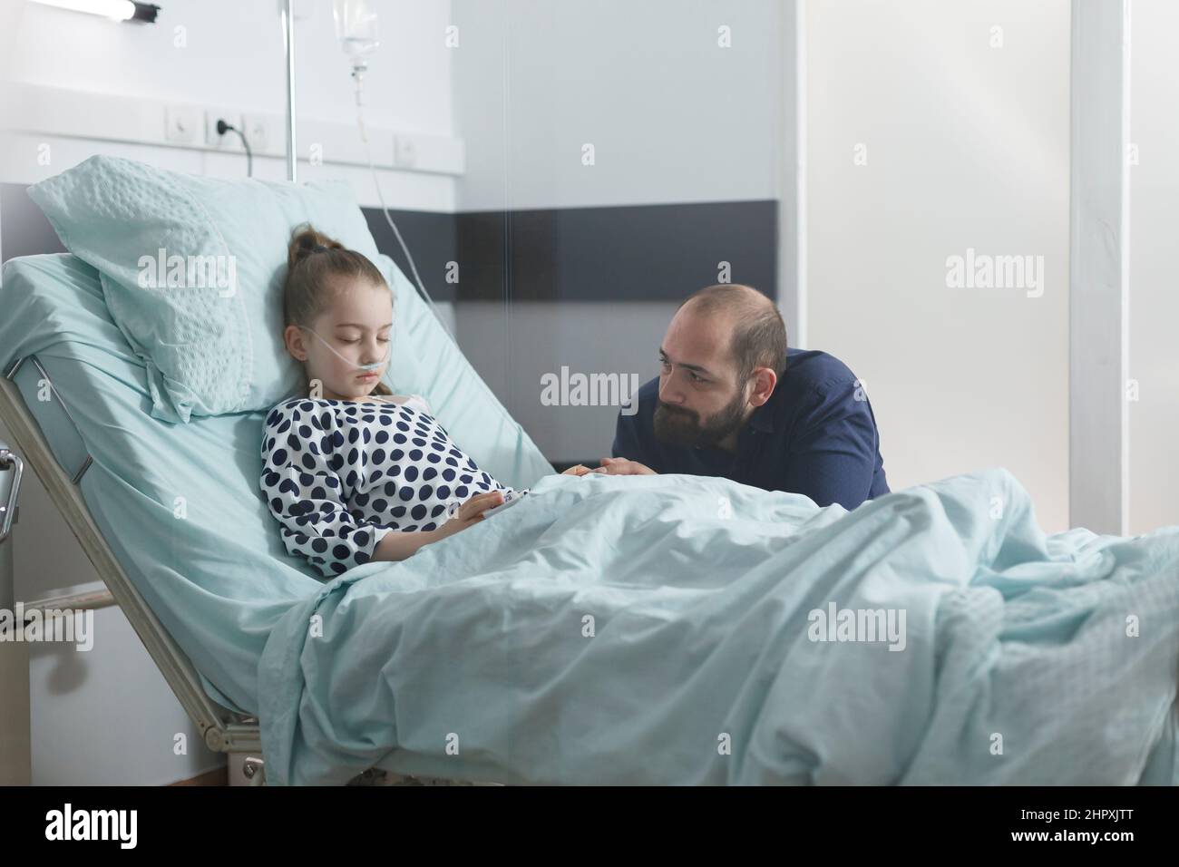 Worried sad uneasy father watching sick little daughter sleeping in hospital bed while in pediatric clinic patient room. Under treatment ill child resting while attentive parent taking care of her. Stock Photo