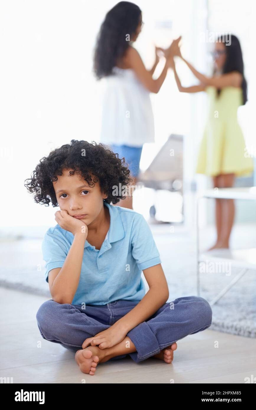 Feeling left out.... Portrait of a sad-looking little boy sitting along with his sisters playing in the background. Stock Photo