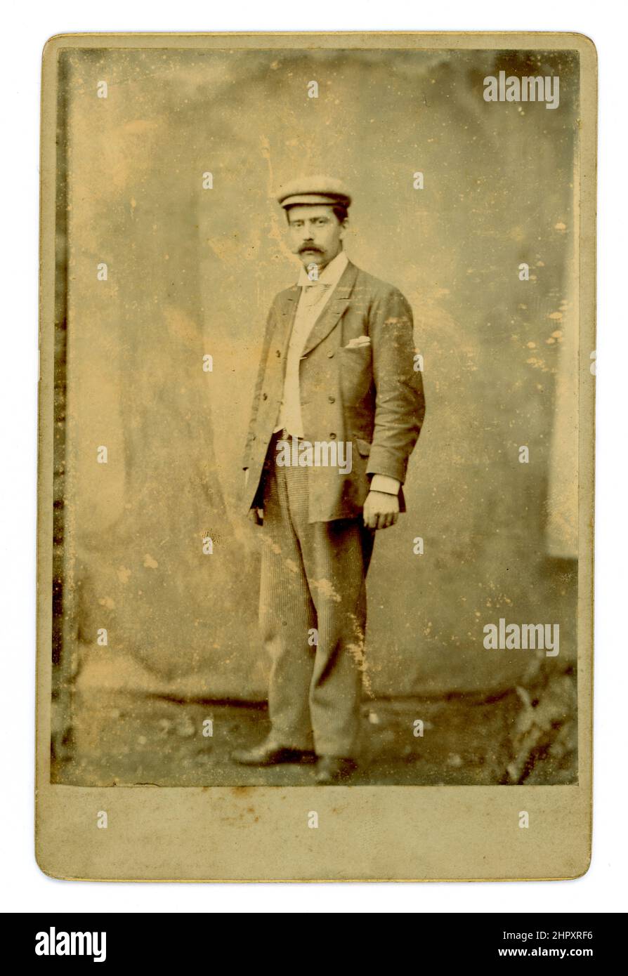 Original Victorian cabinet card portrait of a working class man wearing a flat cap, an ill-fitting suit, with too short sleeves, turned up collar, tie, taken outside with a canvas backdrop against a stone wall - late 1880's, early 1890's, U.K. Stock Photo