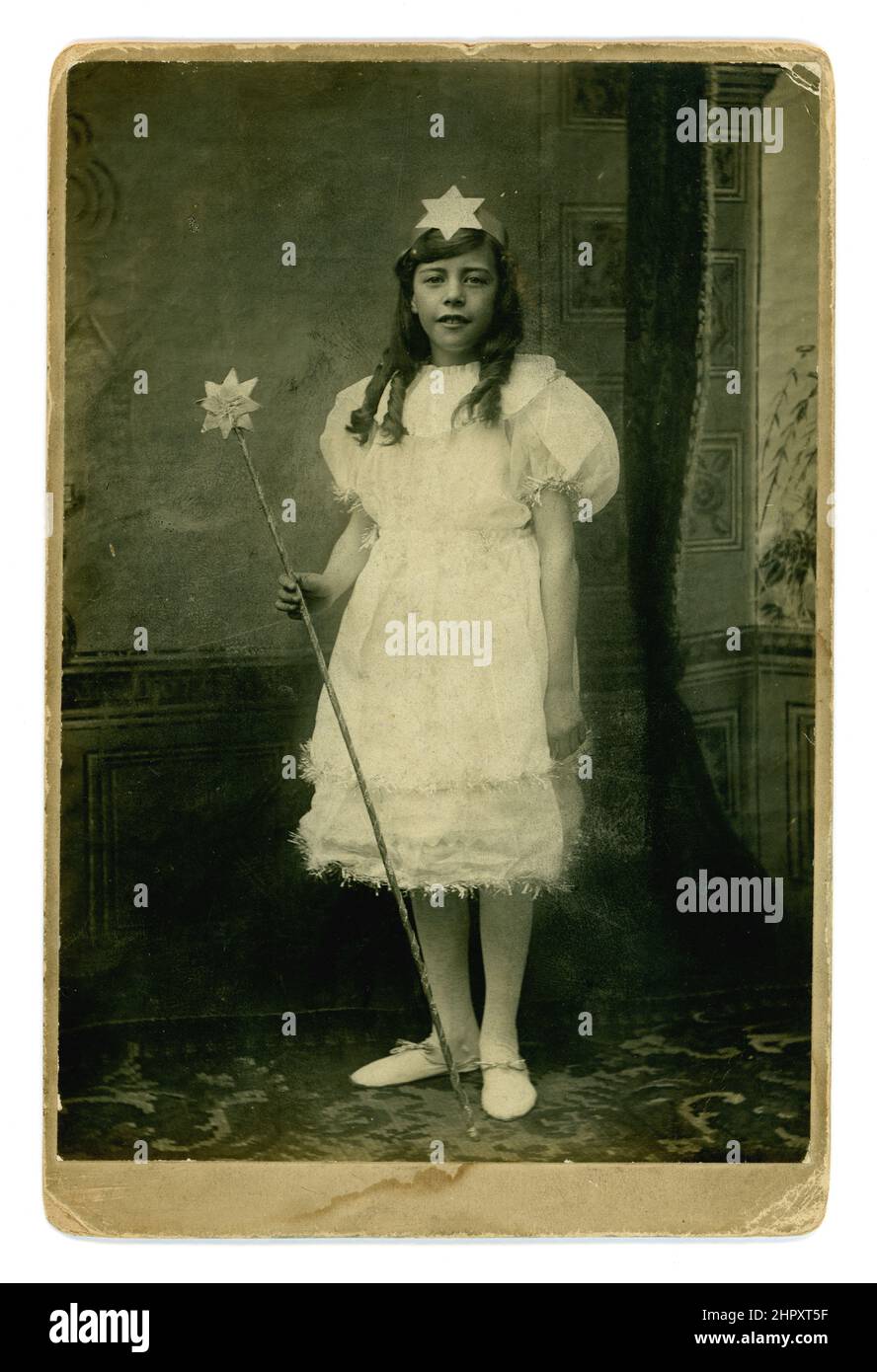 Original and charming Victorian cabinet card studio portrait of smiling older girl wearing a fairy costume with tinsel trim, holding a wand, she has large puff sleeves on a white party dress, fashionable circa 1895. Stock Photo