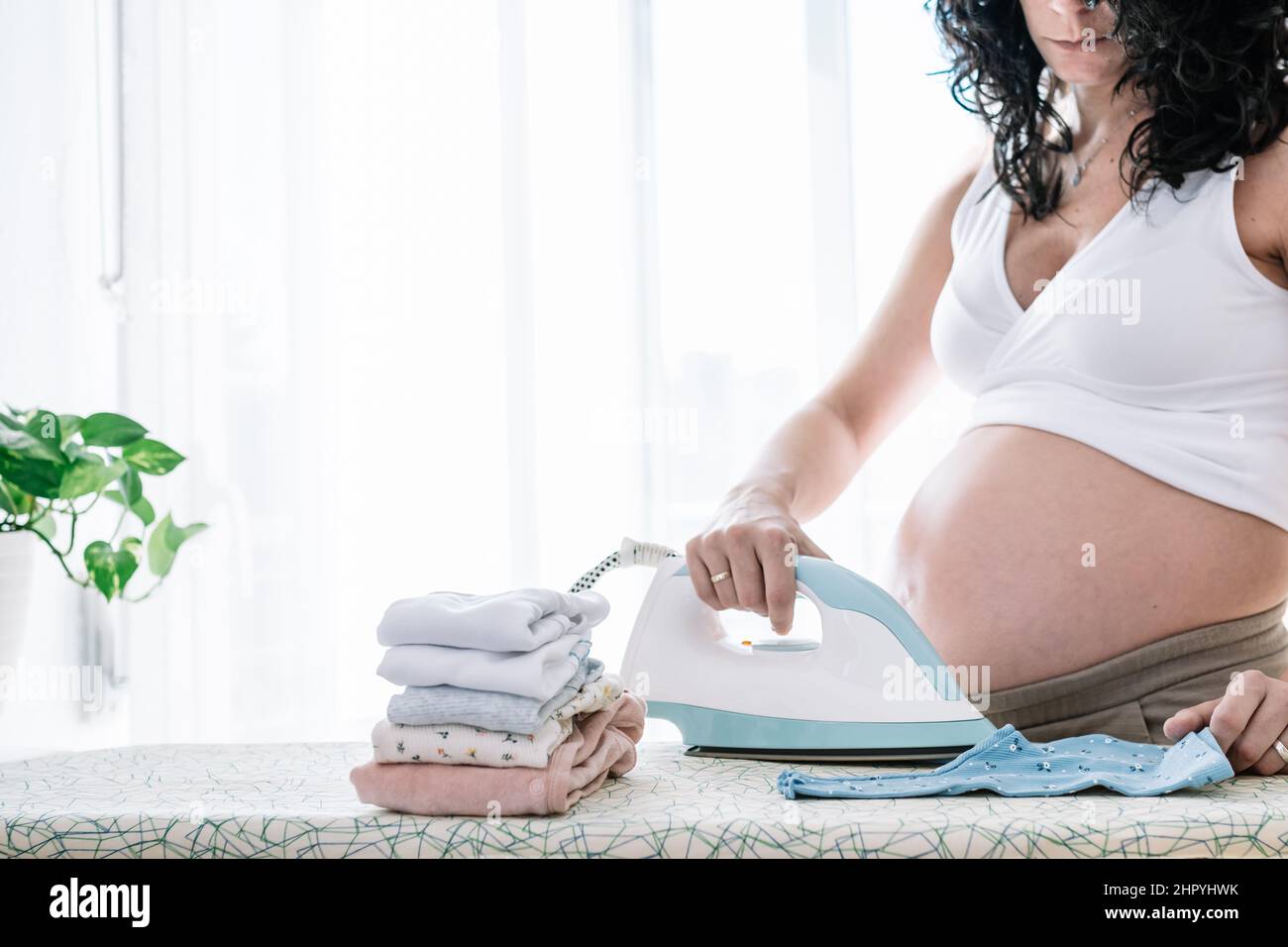 young pretty pregnant woman ironing the clothes of her next baby, preparing the room for the newborn Stock Photo