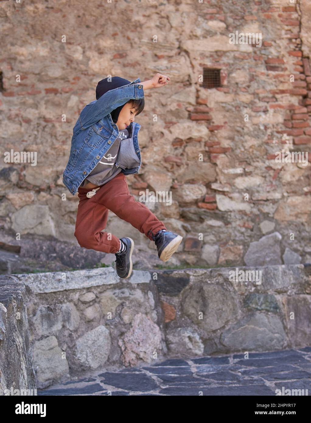 Latin child pirouettes in the air in an old town. Little boy in casual dress with a hooded jean jacket. Concept of parkour in childhood. vertical Stock Photo