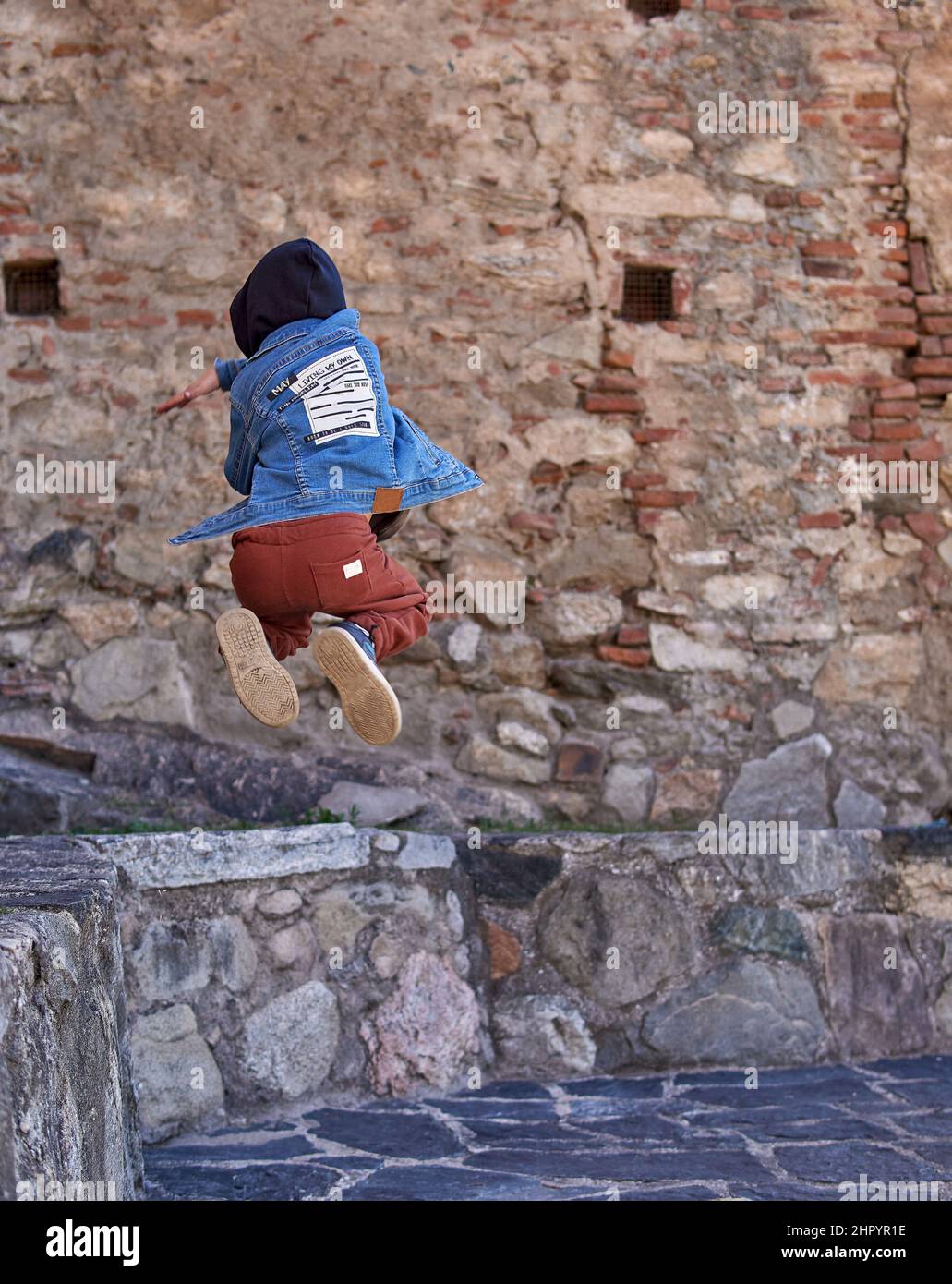 boy Child Practicing Parkour Gymnastics Outside on ancient town. kid on his back in a hooded jean jacket pirouetting on air. Vertical Stock Photo