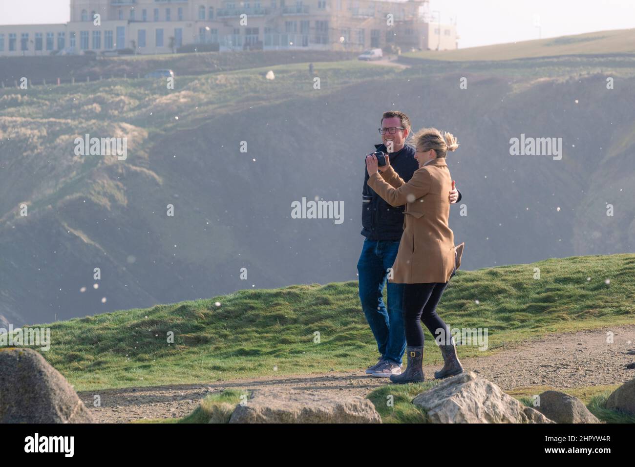 A man supporting his partner as she photographs the wild weather as sea foam blown in the air by storm force wind as Storm Eunice reaches Newquay in C Stock Photo