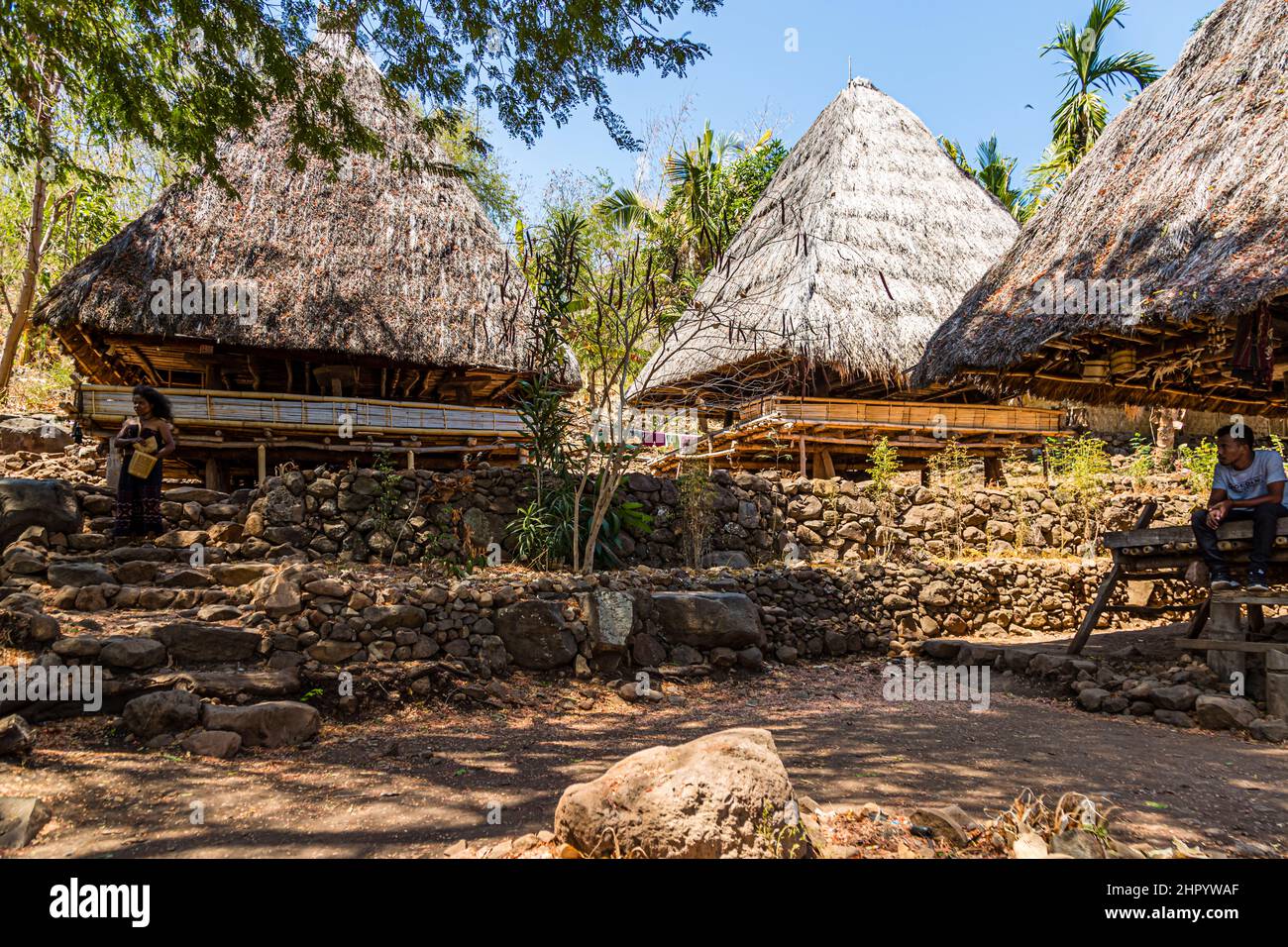 Thatched lumbung storehouses stand on terraced stone walls in the village of Takpala, Alor, Indonesia; a woman stands in front of one of the houses, a man sits on a bench to the right. Stock Photo