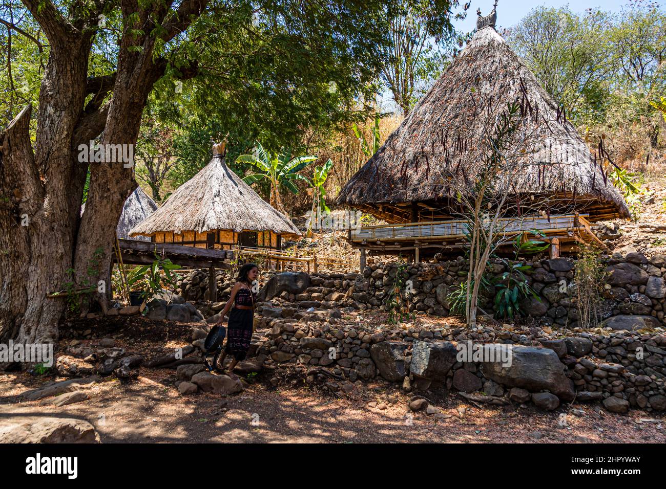 A warrior in a red feathered headdress carries a drum past the lumbung storehouses, with a woman standing by the stone steps in the background, village of Takpala, Alor, Indonesia. Stock Photo