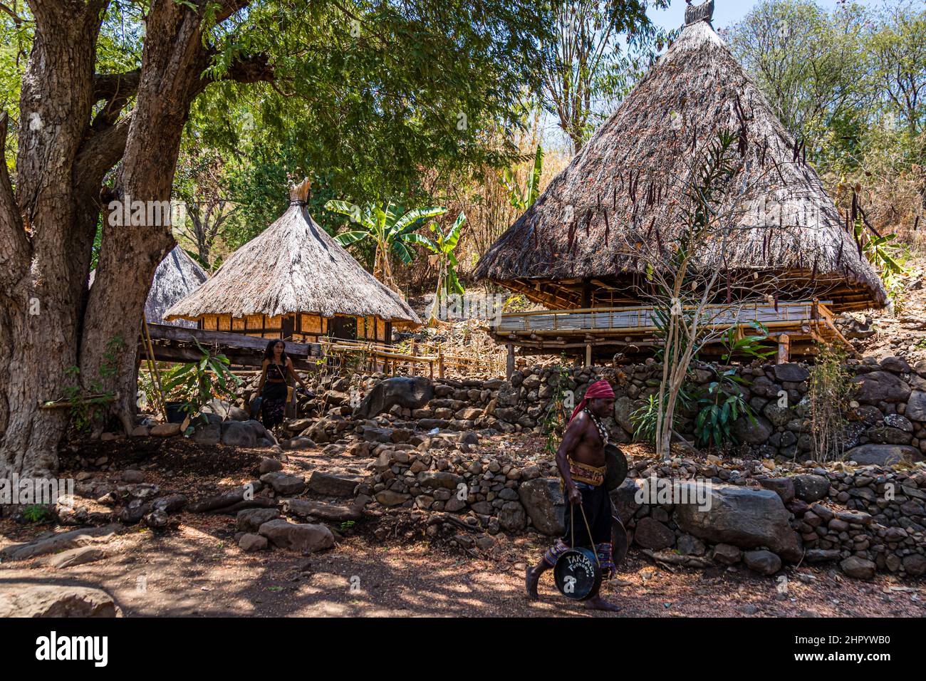 A woman in ikat dress with a red headscarf stands among stone walls in front of two lumbung storehouses in the village of Takpala, Alor, Indonesia. Stock Photo