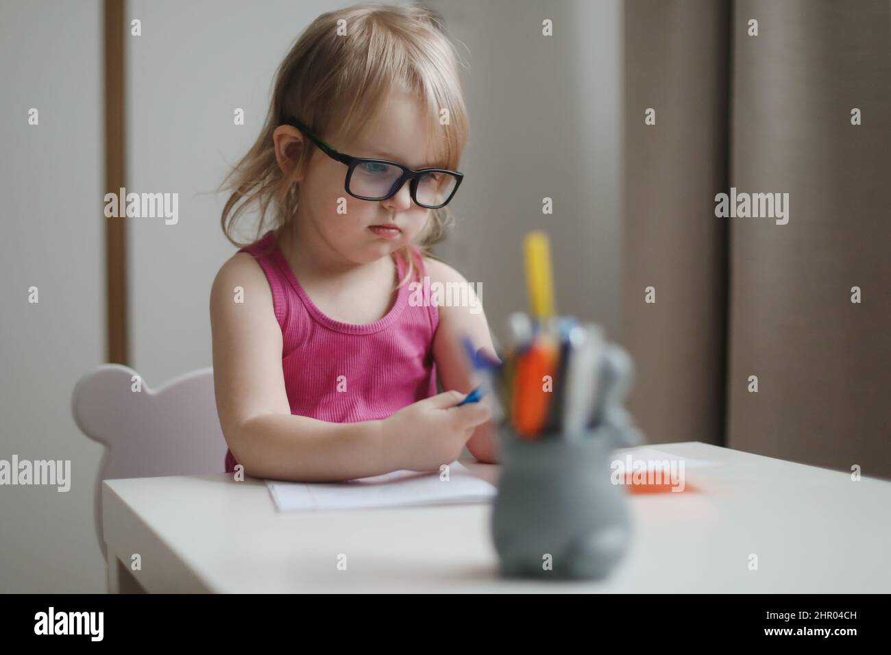 small funny girl in eyeglasses writing and drawing at the table. Stock Photo