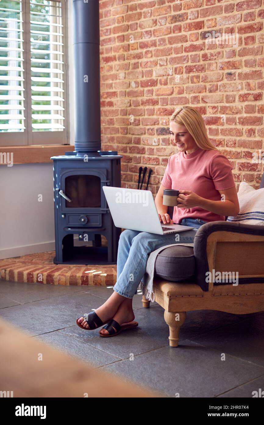 Young Woman Sitting In Lounge Chair Against Exposed Brickwork With Laptop Working From Home Stock Photo