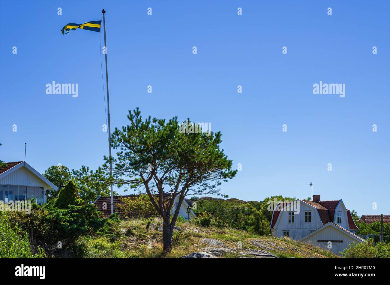 The Swedish national flag flies on a flagpole in the wind on a hill in a settlement of country houses on South Koster Island, Bohuslän, Sweden. Stock Photo