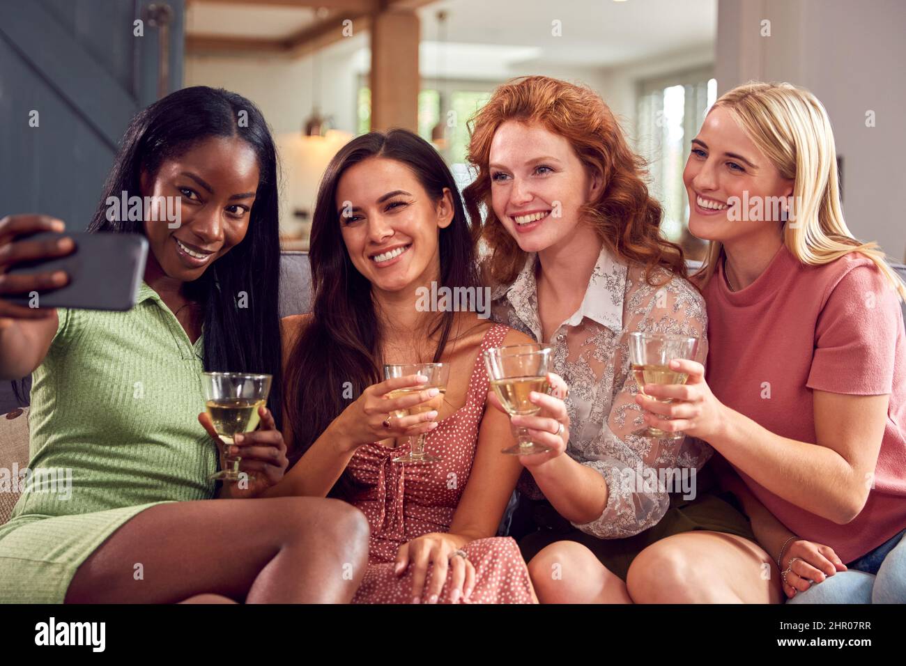 Multi-Cultural Group Of Female Friends Sitting On Sofa At Home Posing For Selfie Together Stock Photo