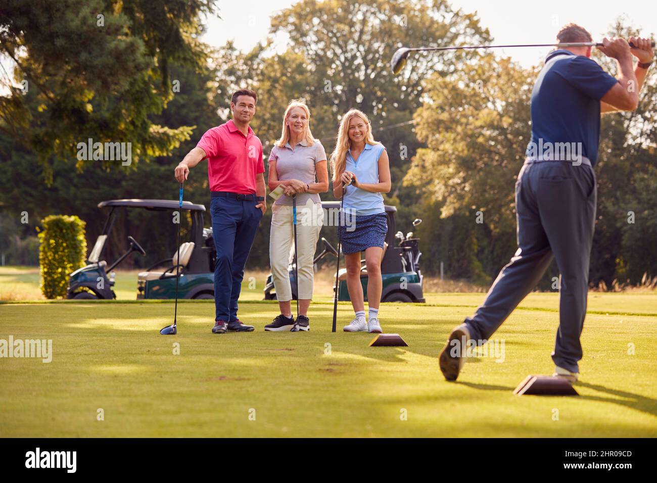 Mature And Mid Adult Couples Standing By Golf Buggy Watching Man Hit Tee Shot Stock Photo