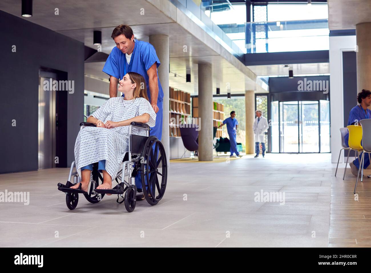 Male Nurse Wearing Scrubs Pushing Female Patient In Wheelchair Through Hospital Building Stock