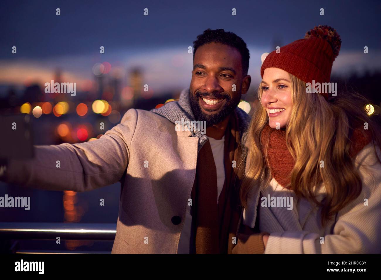 Couple Against City Lights Outdoors Wearing Coats And Scarves Posing For Selfie On Phone At Dusk Stock Photo