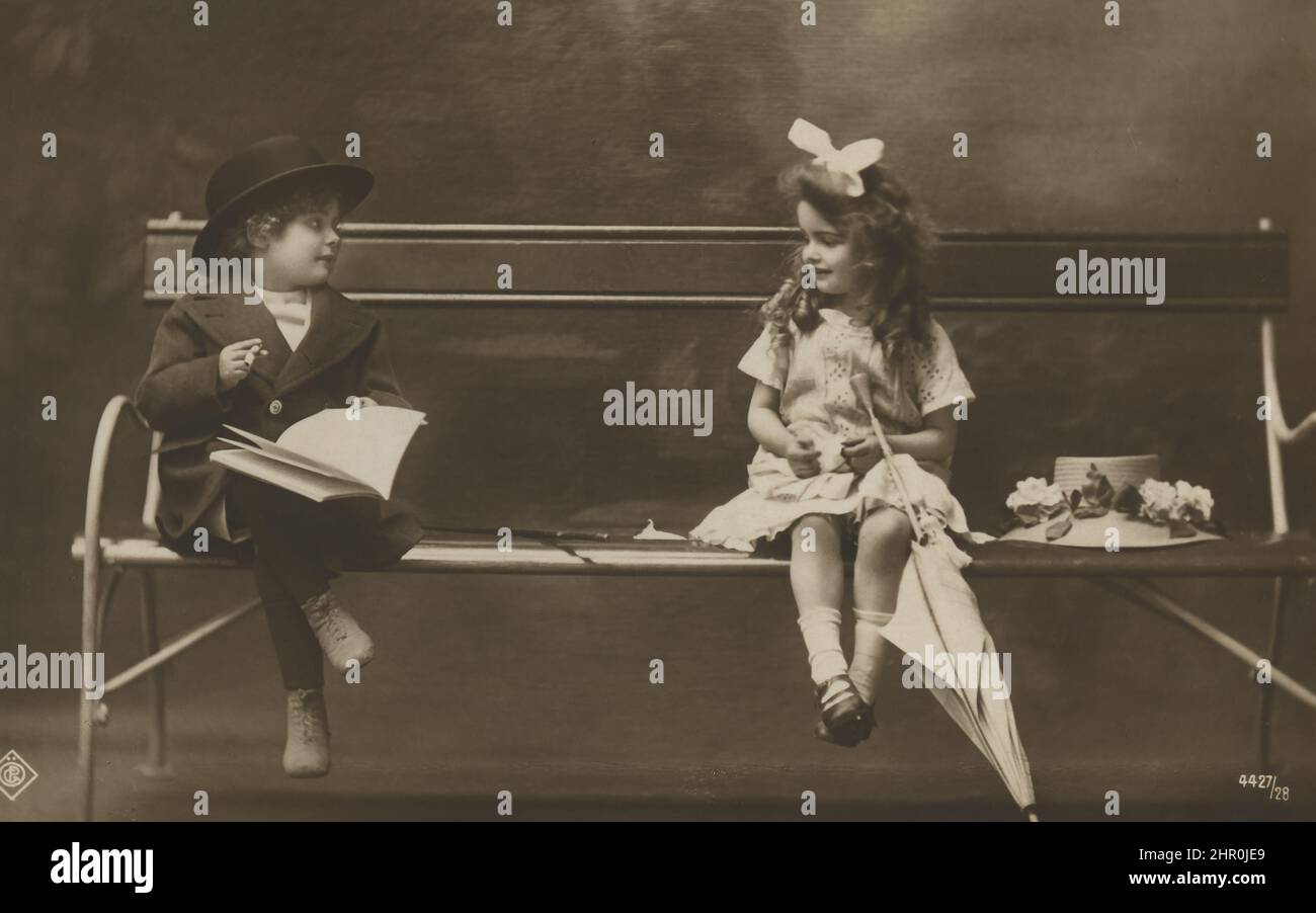 Postcard of children's couple 4-5 years on a bench . Boy in a hat with a book and a cigar. A girl with a big bow in a flirty dress with an umbrella Stock Photo