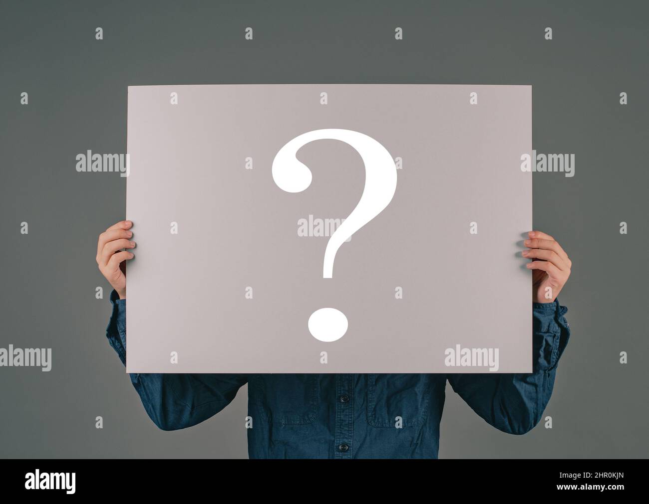 A young man holds a big paper with a question mark, asking why, looking for answers, dark background Stock Photo