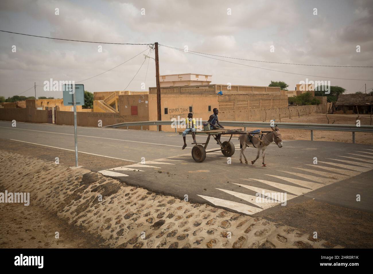 A donkey-drawn cart passes over a speed hump on a modern paved roadway stretching through a small town in northern Senegal, West Africa. Stock Photo