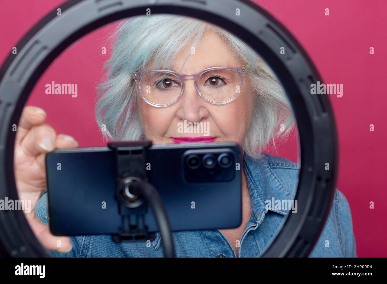 portrait of an older woman with make-up behind a light ring turning on a cell phone to record on pink background Stock Photo
