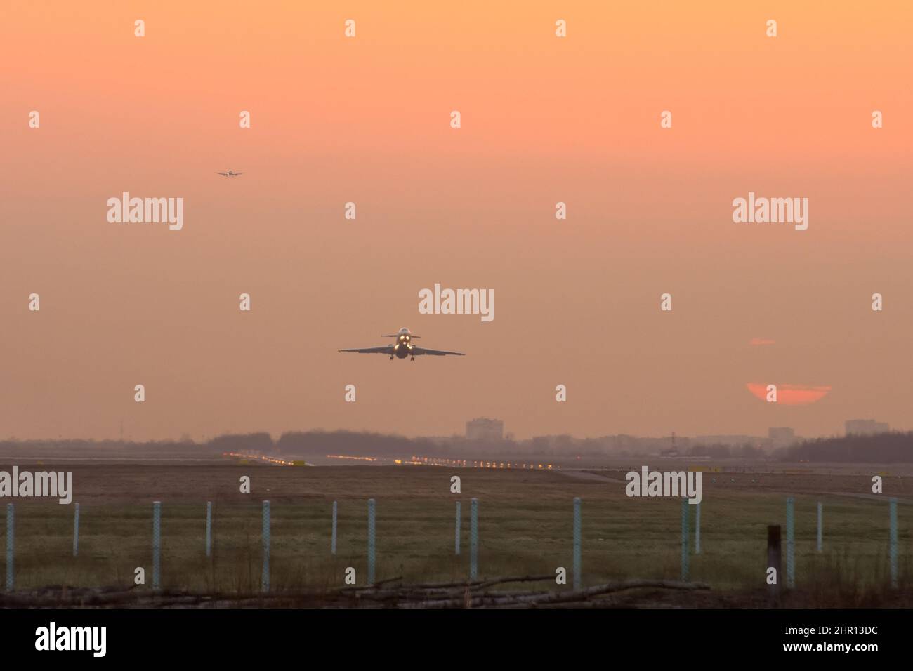 View of the airport and the runway during sunset, drive the sun behind the clouds. One airplane takes off, another in the queue for landing Stock Photo