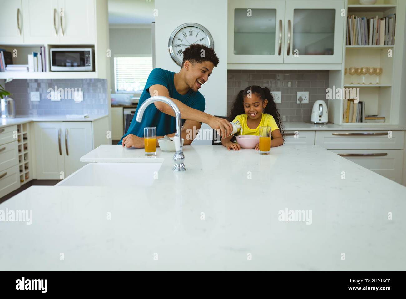 Happy multiracial father pouring milk for daughter sitting at kitchen island during breakfast