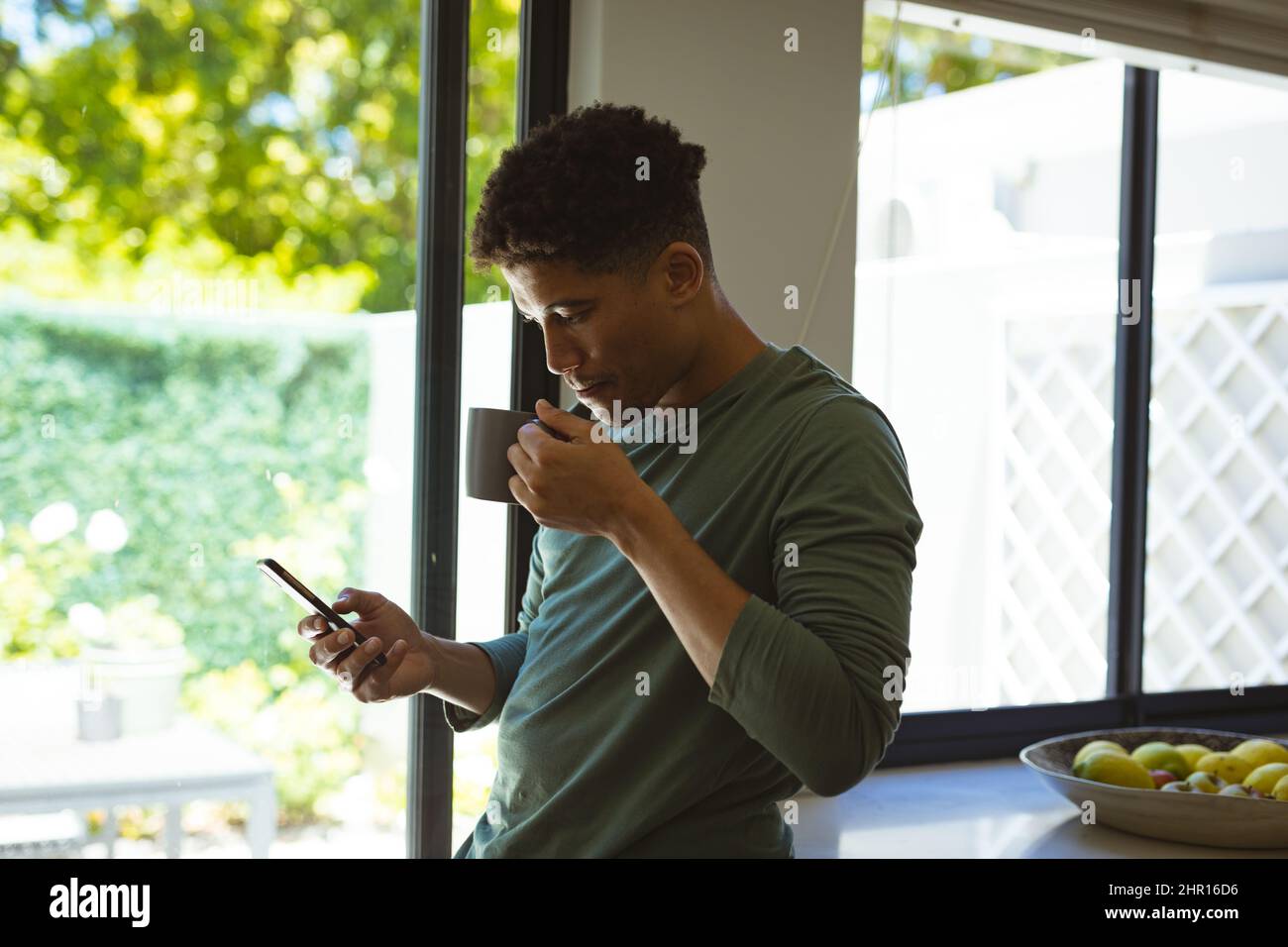 Young biracial man drinking coffee from cup while using smartphone in kitchen at home Stock Photo