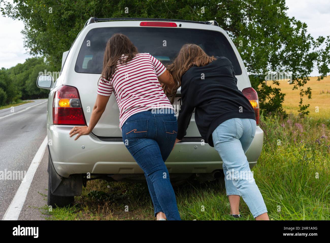 A broken car. Two young women push a broken car on the road, a breakdown, against the background of a yellow field. Stock Photo