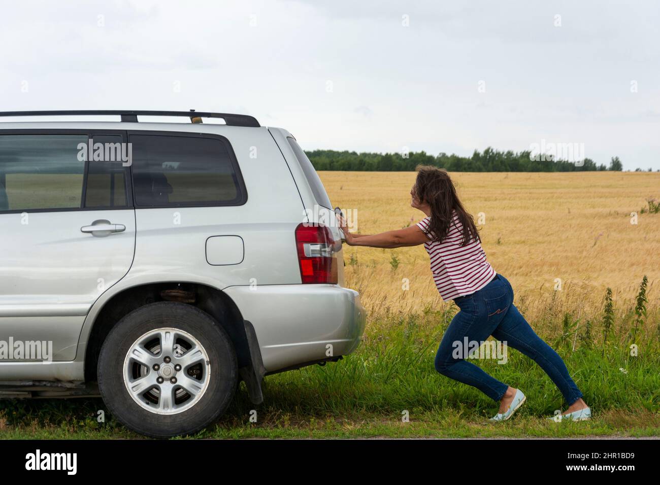 A broken car. A young woman pushes a broken car on the road, a breakdown, against the background of a yellow field. Stock Photo