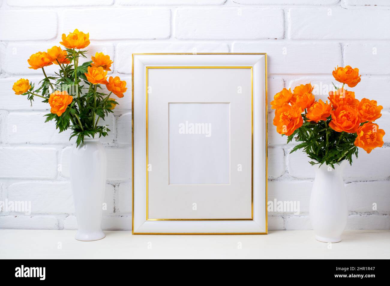 Gold decorated white frame mockup with two vase and globeflowers near painted brick wall. Empty frame mock up for presentation artwork. Template frami Stock Photo