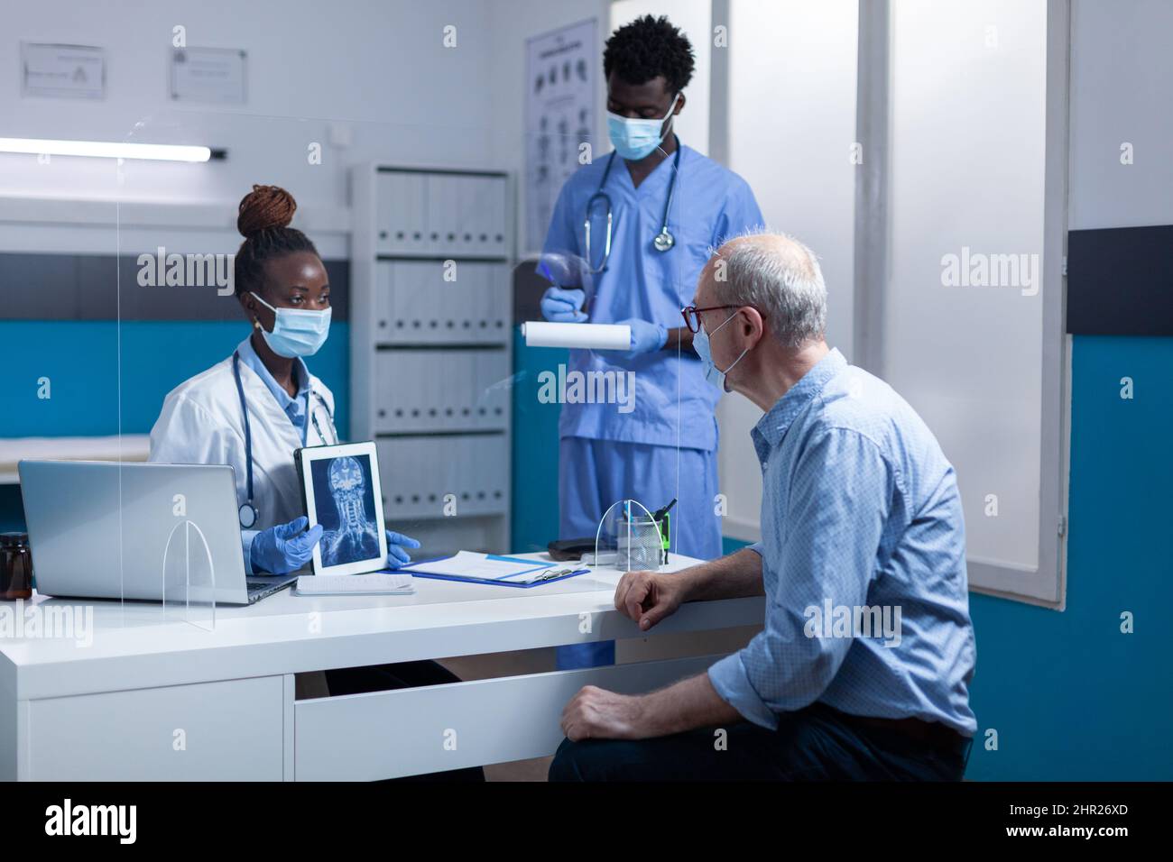Clinic radiology specialist holding x-ray scan image and explaining illness to retired man while medical nurse taking notes on clipboard. Expert radiologist showing MRI scan image to elderly man. Stock Photo