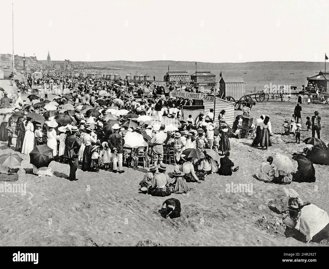 A busy beach scene at the British seaside resort of Weymouth, Dorset, England, UK c. 1900. Seaside entertainment on a wooden stage set up on the beach features a troupe called ‘The Lyric Concert Party’. They wear white trousers and straw boater and sailor’s hats. The audience stand or sit mainly on wooden chairs – many with umbrellas and parasols to protect from the sun. Some children play in the sand. In the background are bathing huts and wheeled bathing machines. One large cabin has a star on its roof and a sign indicating that it is a ‘Gentleman’s Saloon’ – a vintage Victorian photograph. Stock Photo
