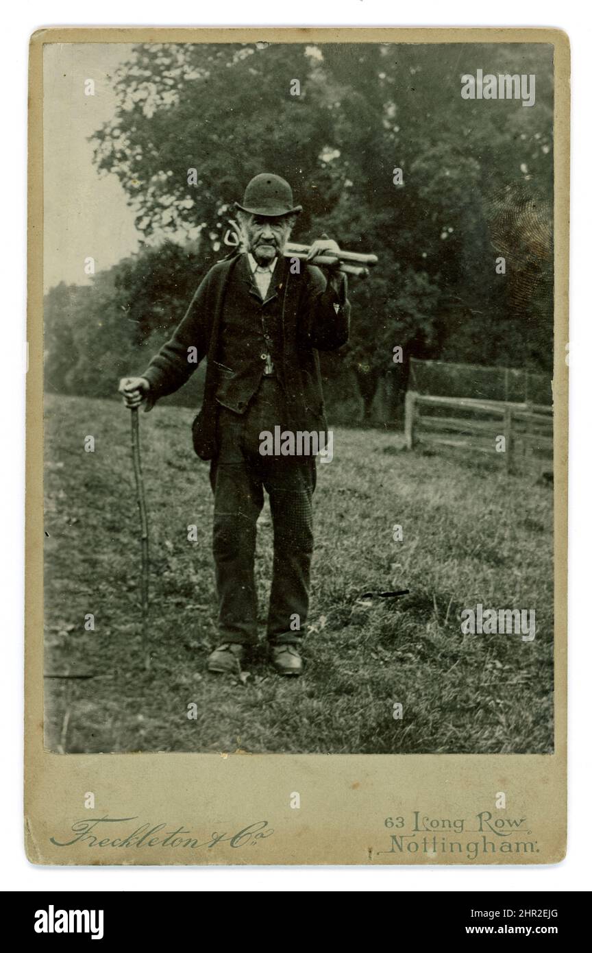 Original and rare Victorian cabinet card of poor working class older farm labourer, wearing his patched working suit, a bowler hat typical of agricultural labourers of the time, carrying farm implements, possibly hoes, rakes or billhooks. HIs looks a hard life -he is of pensioner's age. Photograph by Freckleton & Co., 63 Long Row, Nottingham, U.K. circa 1890's. Stock Photo