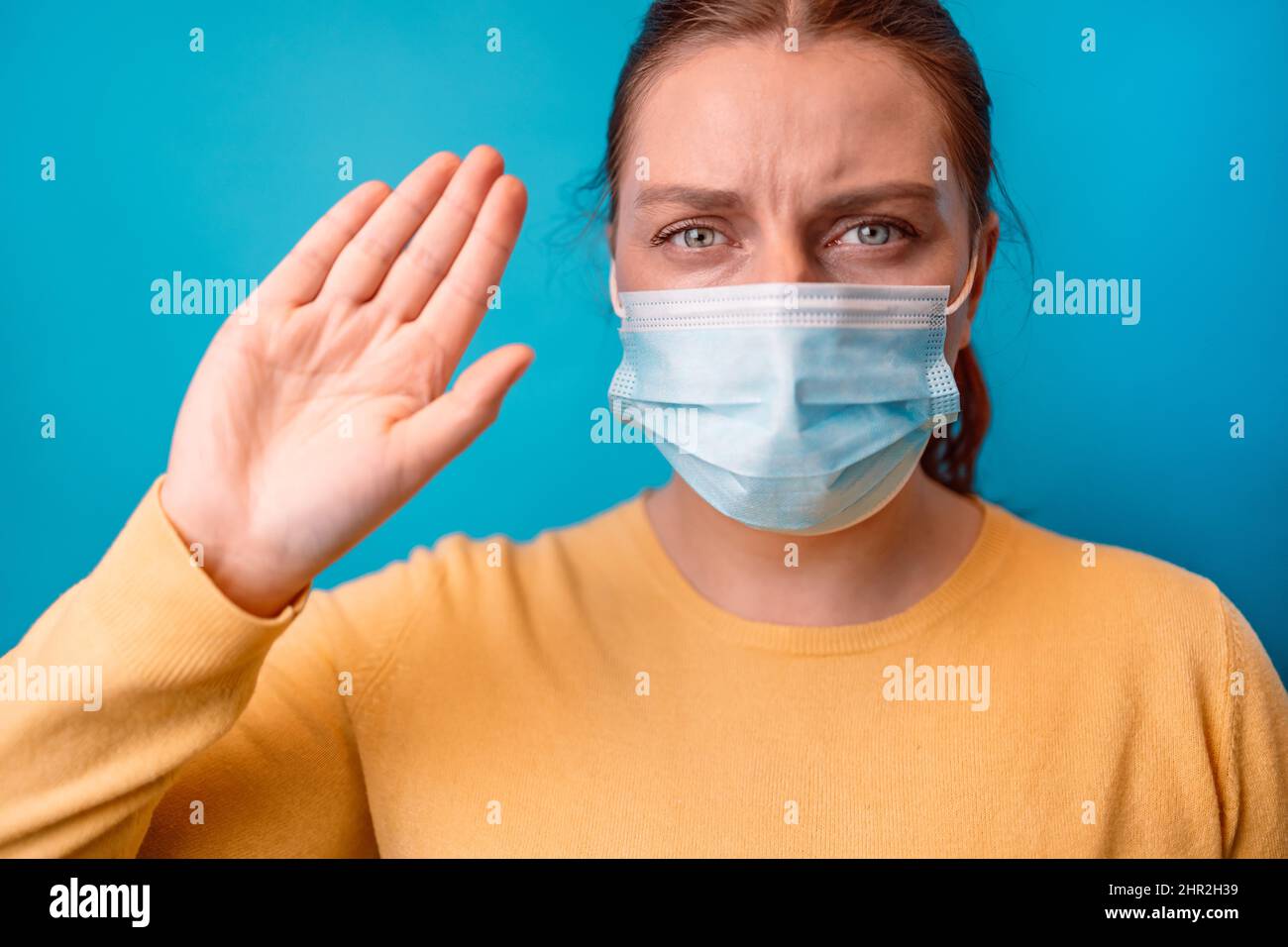 Young woman in a protective medical mask with yellow sweater showing stop gesture over isolated blue background Stock Photo