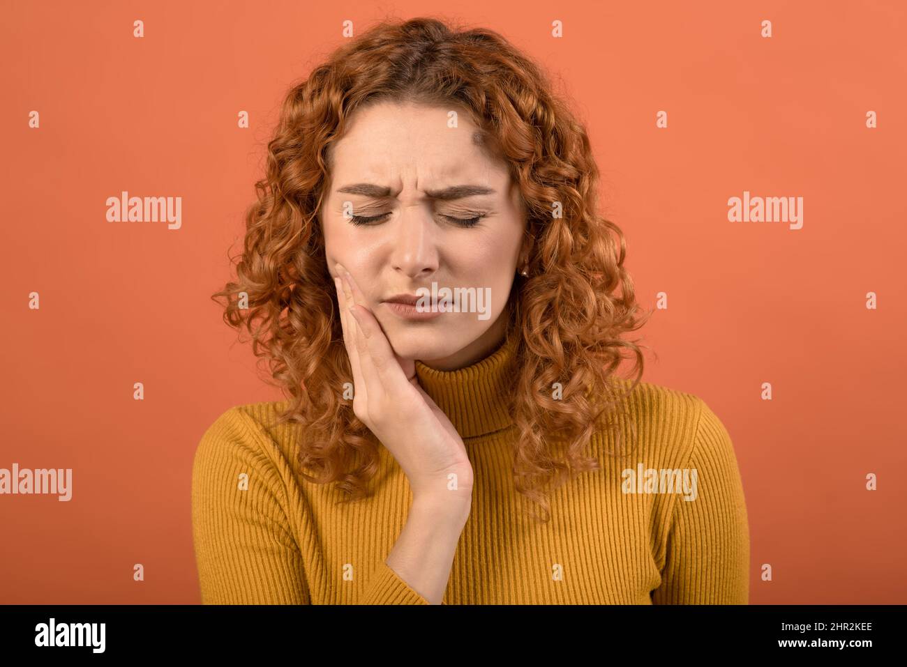Young and attractive caucasian redhead girl touching her mouth with toothache isolated on orange studio background. Stock Photo