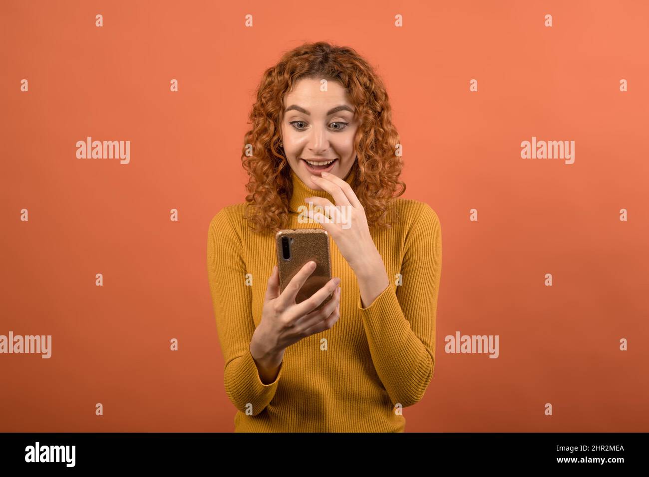 A young and attractive redhead Caucasian girl in an orange jumper using a smartphone and the girl is shocked isolated on an orange studio background. Stock Photo